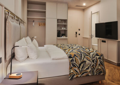 doppelbett-bunte-tagesdecke-schrank-mit-kleiderbuegel-tv-offene-tuer-vom-badezimmer