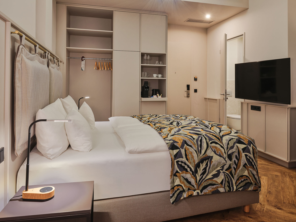 doppelbett-bunte-tagesdecke-schrank-mit-kleiderbuegel-tv-offene-tuer-vom-badezimmer