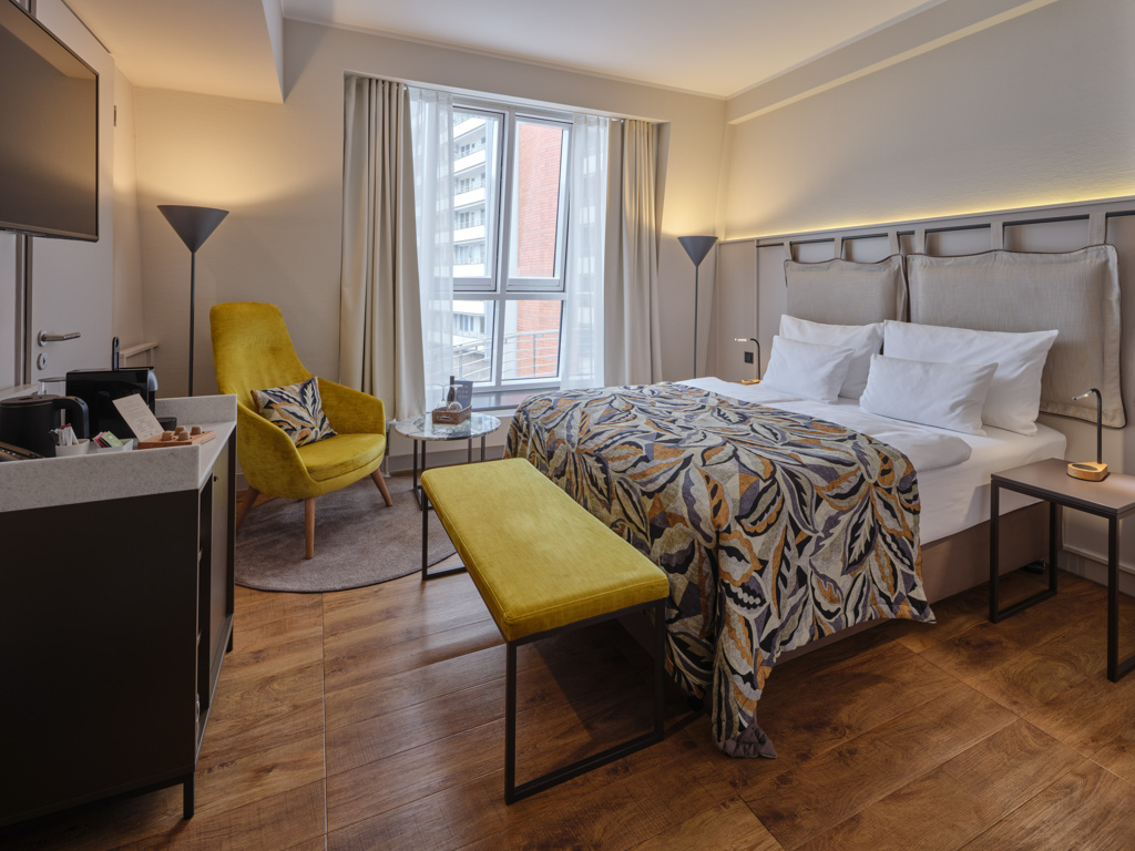 doppelbett-bunte-tagesdecke-gelbe-fußbank-gelber-sessel-seitliches-fenster